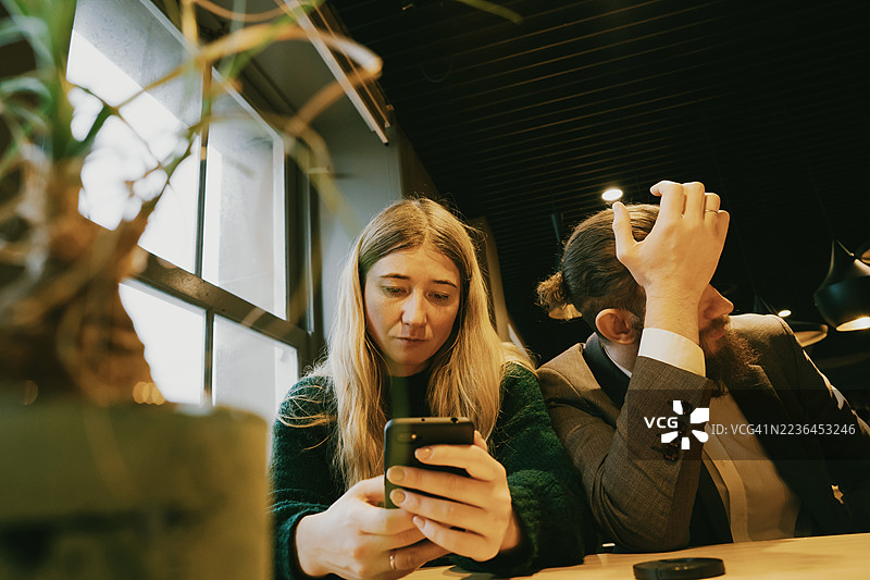 两位同事在咖啡馆，一人专注于手机，另一人看起来紧张而沉思。图片素材