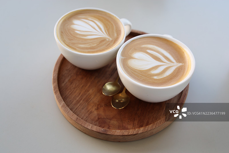 两杯白色杯子的平白咖啡放在圆形木质托盘上，旁边有金色茶匙。图片素材