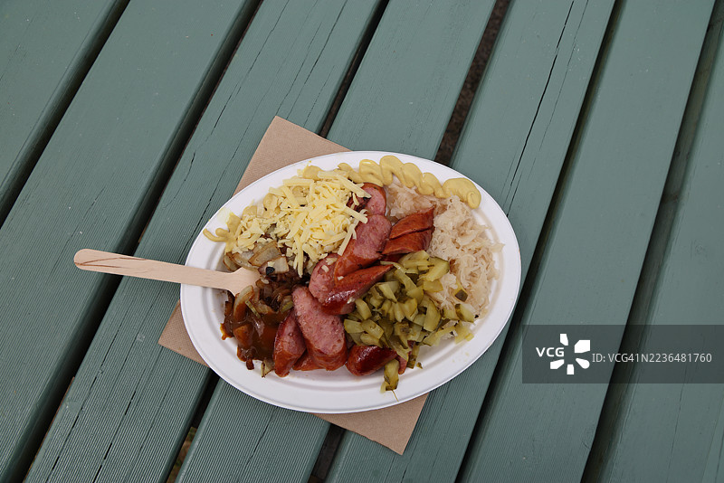 切片熏制香肠、炸洋葱和土豆、切碎的黄瓜腌菜、酸菜、德国咖喱番茄酱、磨碎的奶酪和芥末，放在纸板盘上，配有一次性木叉和纸巾，摆放在野餐桌上。图片素材