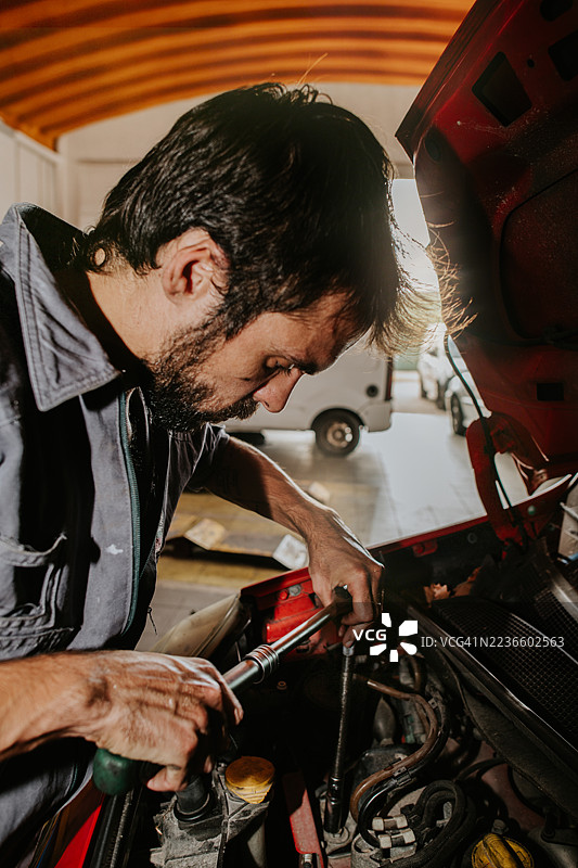 一名穿着工装的机械师在红色汽车的引擎盖下使用扳手。图片素材