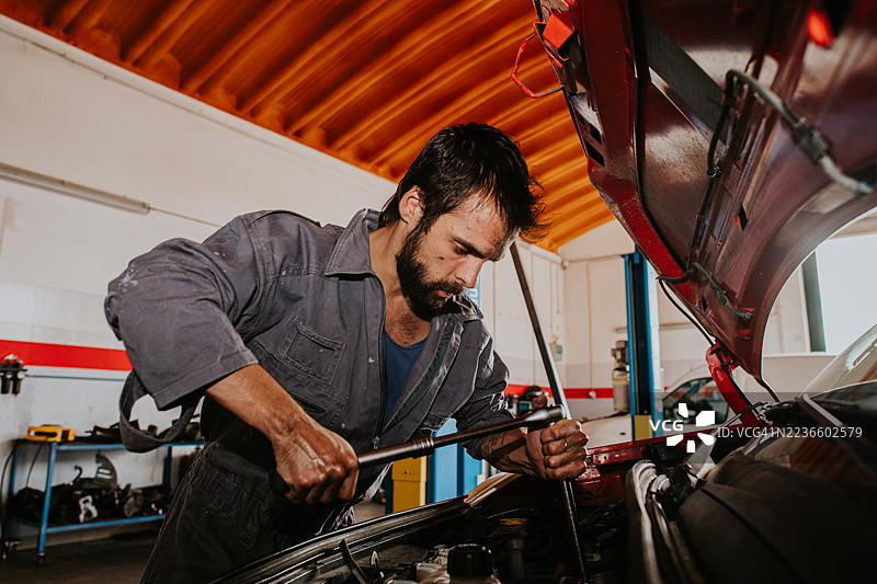 一名穿着工装的机械师在红色汽车的引擎盖下使用扳手。图片素材