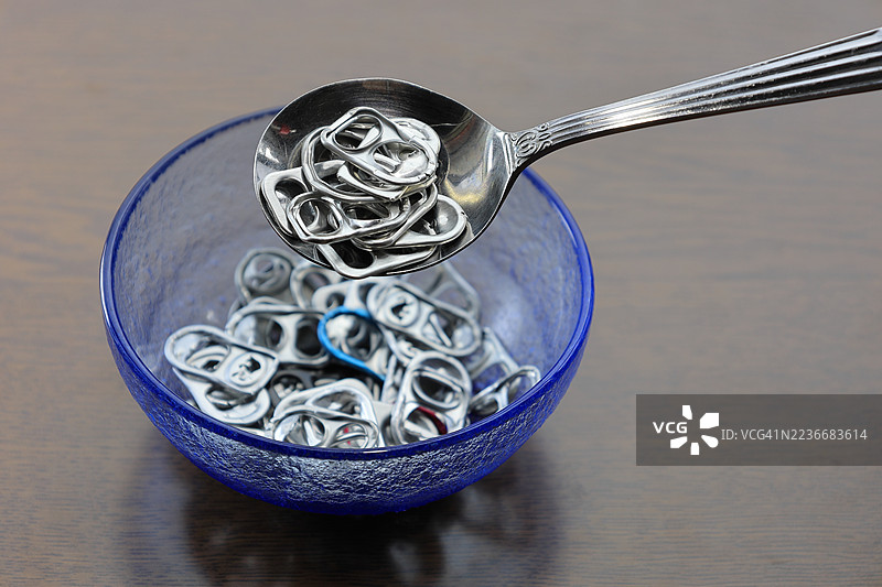 一个特写镜头，勺子从木桌上的蓝色玻璃碗中舀起铝罐拉环。拉环是银色的，还有一些是红色、蓝色和金色的。这个画面创意地像是碗中的谷物，象征着回收和环保意识。图片素材