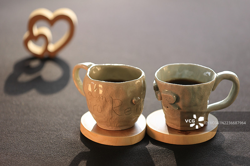 一个温暖而迷人的场景，两个手工陶瓷杯放在圆形木质杯垫上。杯子具有乡村风格的手工质感，一个上面刻有文字，另一个则压印有字母“R”。两个杯子都装满了咖啡。图片素材