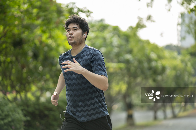 一名男子在户外慢跑，身穿蓝色锯齿形图案T恤，背景为绿色植被。表情专注而坚定。图片素材
