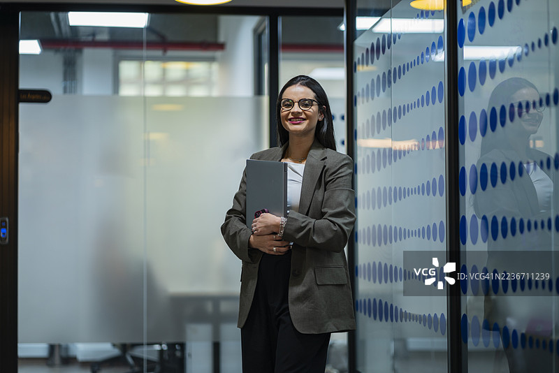 一位女商人在办公室走廊里边走边看，她戴着眼镜，手里拿着笔记本电脑和手机。图片素材