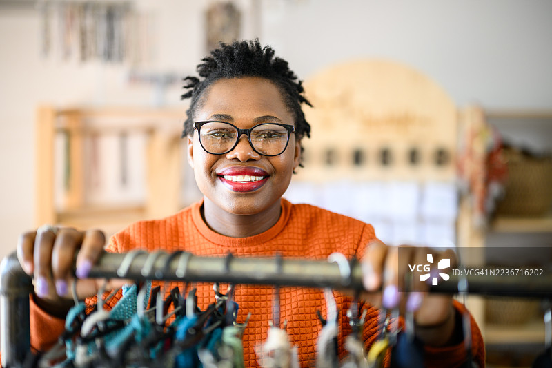 特写肖像，一位面带微笑的女性站在摆满色彩鲜艳的手工服装的衣架后面。图片素材
