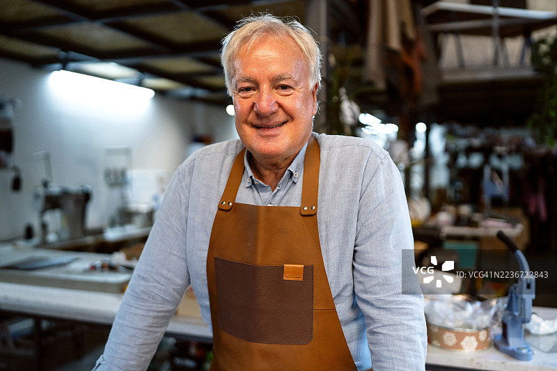 在他的皮革工作室里，微笑自豪的老工匠肖像，周围是工具和材料。图片素材