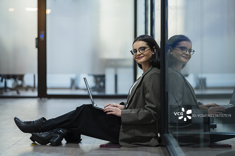 一位女士戴着眼镜，手里拿着笔记本电脑和手机，坐在办公室走廊里。图片素材