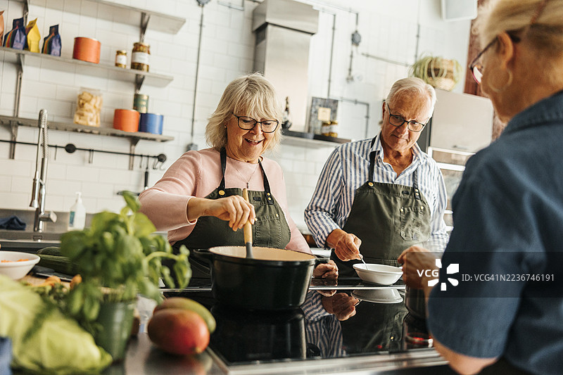 快乐的老年女性在商业厨房的烹饪课堂上与朋友们一起学习，边搅拌锅里的食物。图片素材