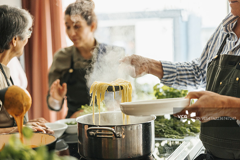 老年男子在商业厨房的烹饪课堂上用漏网从锅中取出意大利面。图片素材