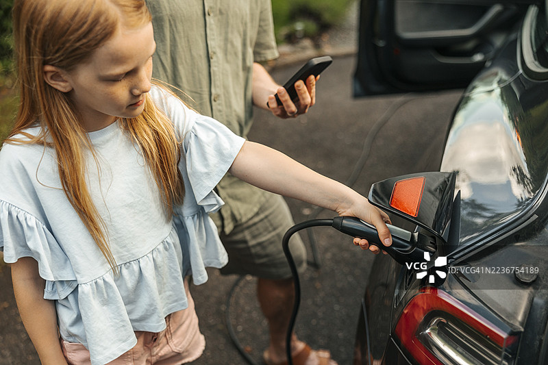 女孩在父亲身边站着，从高角度视角充电电动车插头。图片素材
