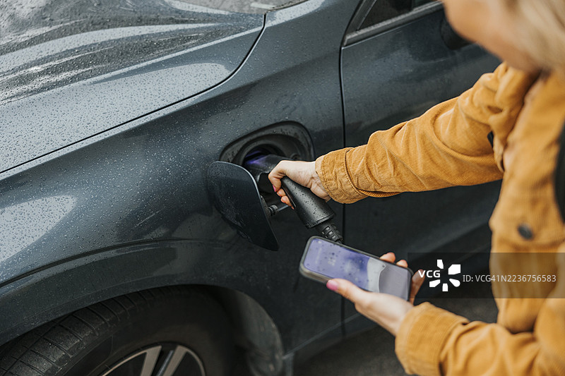 高角度视图的女性在为电动车充电时使用智能手机图片素材