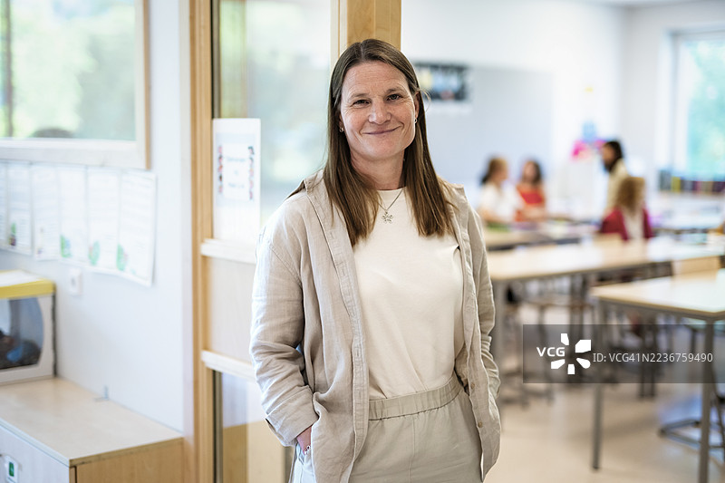 Portrait of smiling female teacher standing with hands in pockets at classroom doorway图片素材