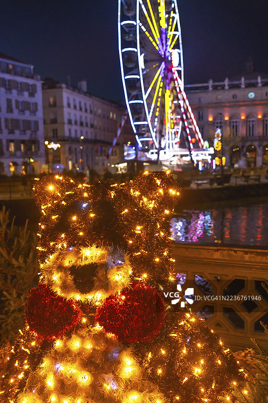 在法国巴约讷，发光的圣诞熊装饰在夜晚闪耀，与摩天轮和节日灯光照亮的背景相映成趣。图片素材