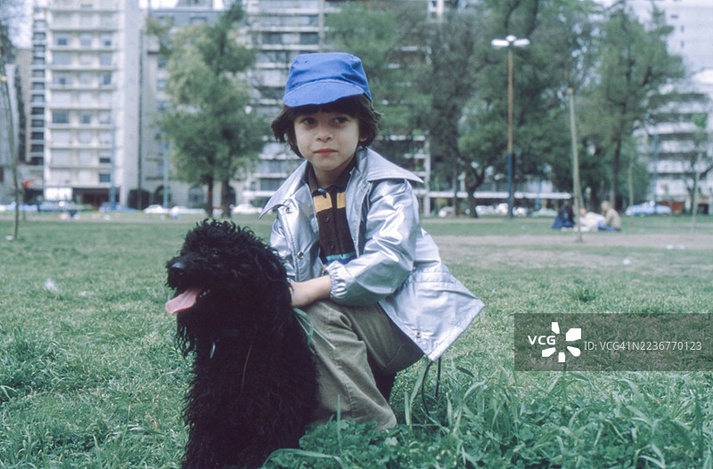 1980年，布宜诺斯艾利斯，一位6岁的小男孩穿着银色夹克和他心爱的贵宾犬在公园里的复古照片图片素材