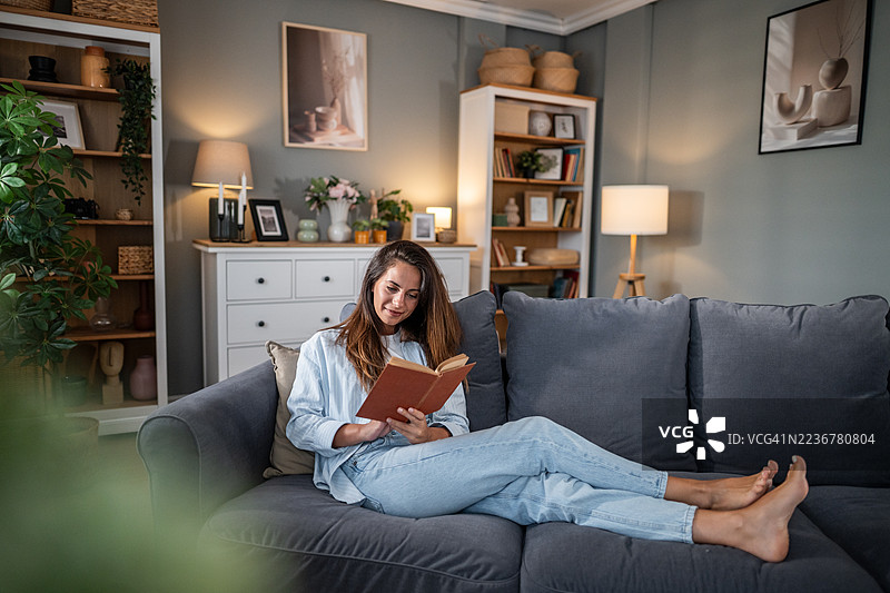 放松的年轻女性在舒适的现代客厅里阅读书籍图片素材