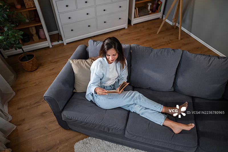 放松的年轻女性在舒适的灰色沙发上阅读书籍图片素材