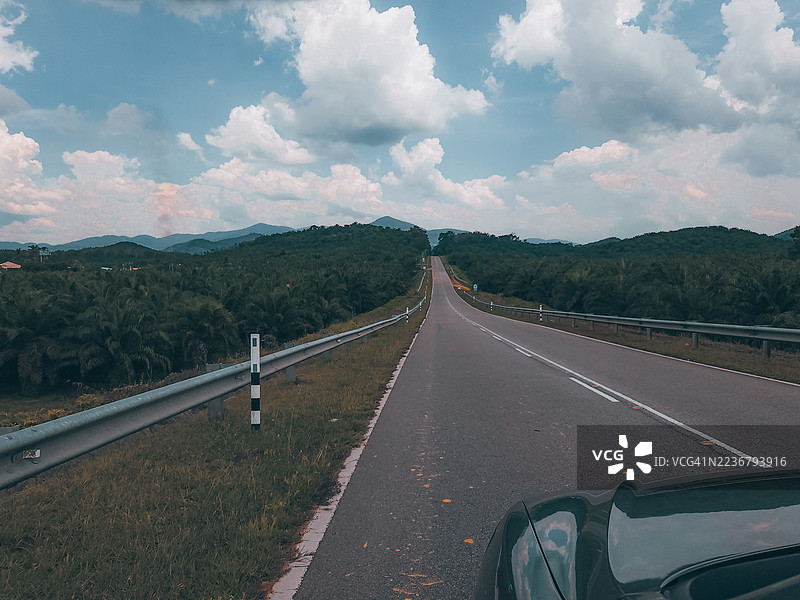一条漫长的空旷道路延伸至远方，穿过郁郁葱葱的绿色景观，天空多云，前景中可见一辆汽车，位于马来西亚的Pertang。图片素材