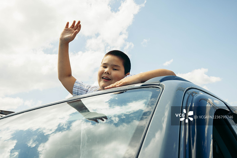 快乐的男孩在车窗外挥手，背景是明亮的蓝天和白云，享受着旅程。图片素材