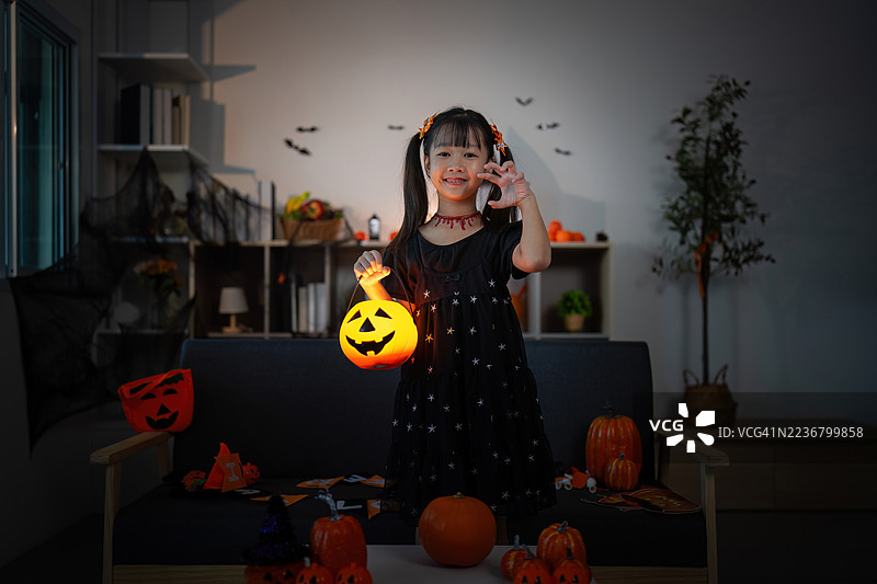 微笑的女孩穿着万圣节服装，手里拿着南瓜灯，站在装饰好的客厅里图片素材