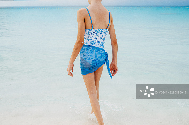 一个穿着蓝色泳衣的十几岁女孩，走在巴哈马海滩边缘清澈浅水区。图片素材