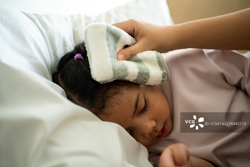 亚洲母亲在家中床上为生病的女孩擦额头以缓解发烧，父母关心孩子的健康保险和家庭医疗保障。图片素材