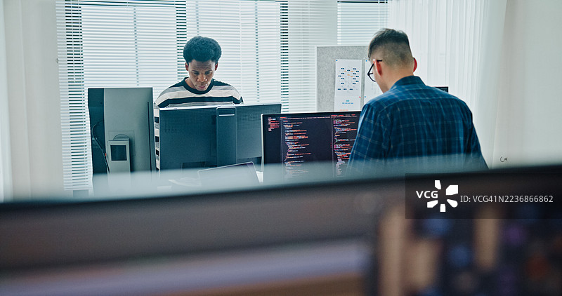 两名男性软件开发人员在办公室的桌子旁站着，分析数据编码并在多个屏幕上工作。人工智能与编程。图片素材
