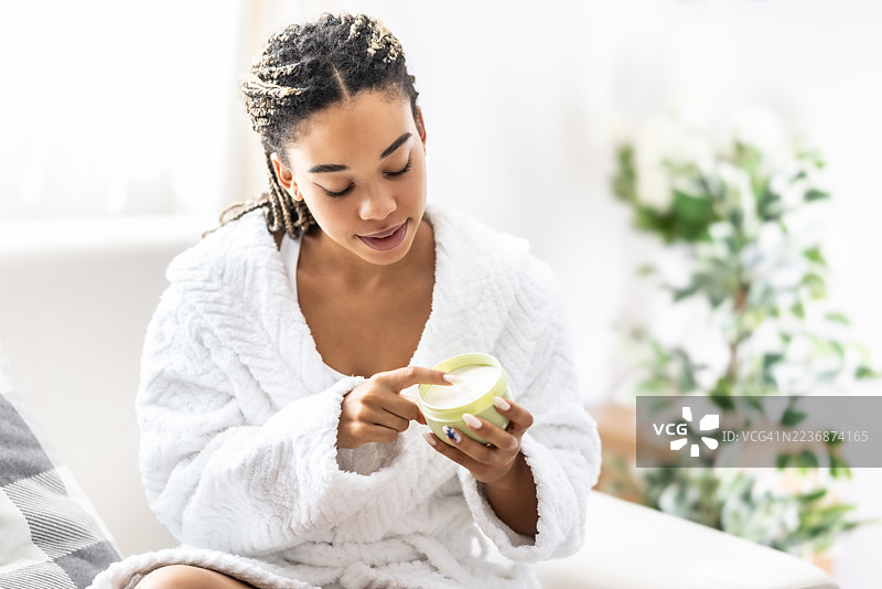 一位穿着白色浴袍的年轻女性手里拿着一个打开的白色奶油容器。她用右手的食指触碰容器中的奶油。图片素材