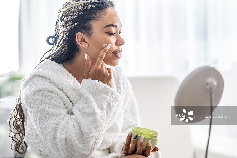 年轻女性穿着白色浴袍，手持一瓶面霜，正在涂抹在脸上图片素材