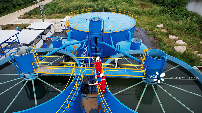 两名穿着红色工作服和个人防护装备的工业工人或工程师走上楼梯，准备在大型水箱的顶部进行工作，水处理设施，市政水处理厂。图片素材