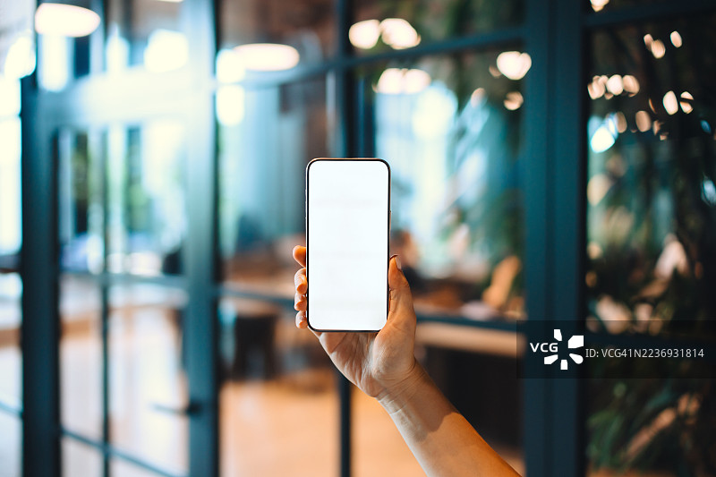 女性手握着空白屏幕的智能手机，背景是现代办公大楼。商业生活方式与科技。银行、金融和投资。智能移动银行。金融科技。设计模板的空白屏幕。图片素材