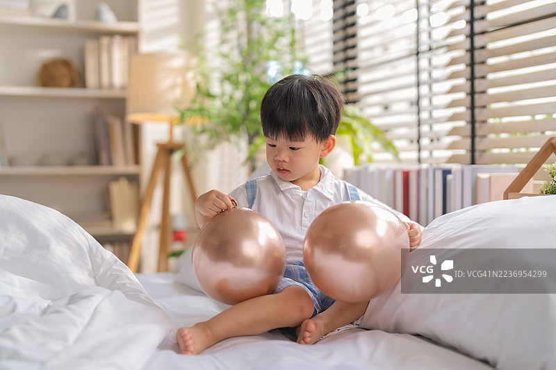 可爱的亚洲幼儿男孩在家中玩耍，手里拿着五颜六色的气球，开心地笑着，享受着快乐的周末早晨活动，展现幸福的童年生活瞬间。图片素材