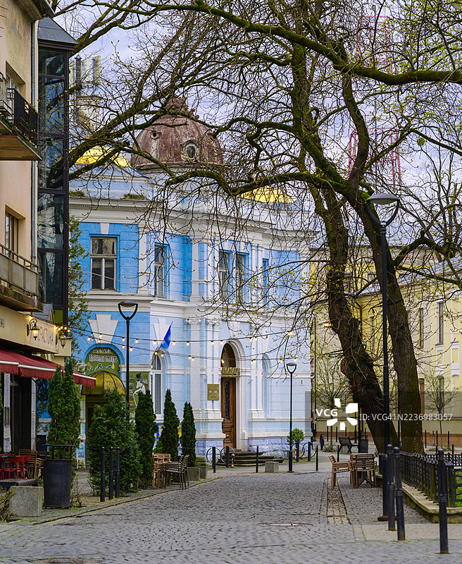 乌克兰伊万诺-弗兰基夫斯克的鹅卵石街道，特色是带圆顶的历史蓝色建筑和早春光秃树下的户外咖啡区图片素材