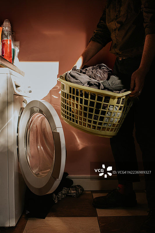 一名男子在洗衣房里拿着洗衣篮，准备将其放入现代洗衣机中。图片素材