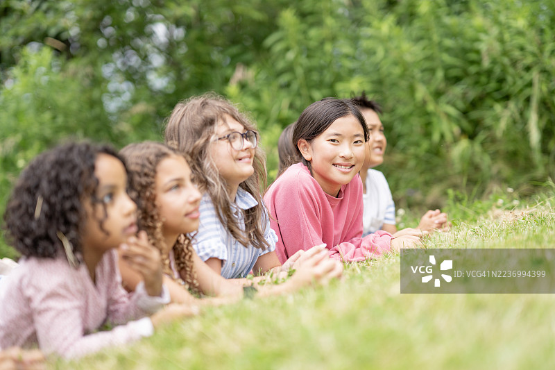 一群多样化的孩子躺在户外的草地上，微笑着观看自然中的某些事物。图片素材
