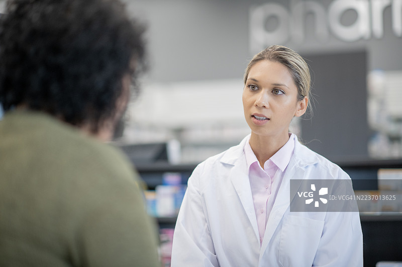 穿着白色实验室外套的专业女性药剂师在药店与顾客讨论药物。图片素材