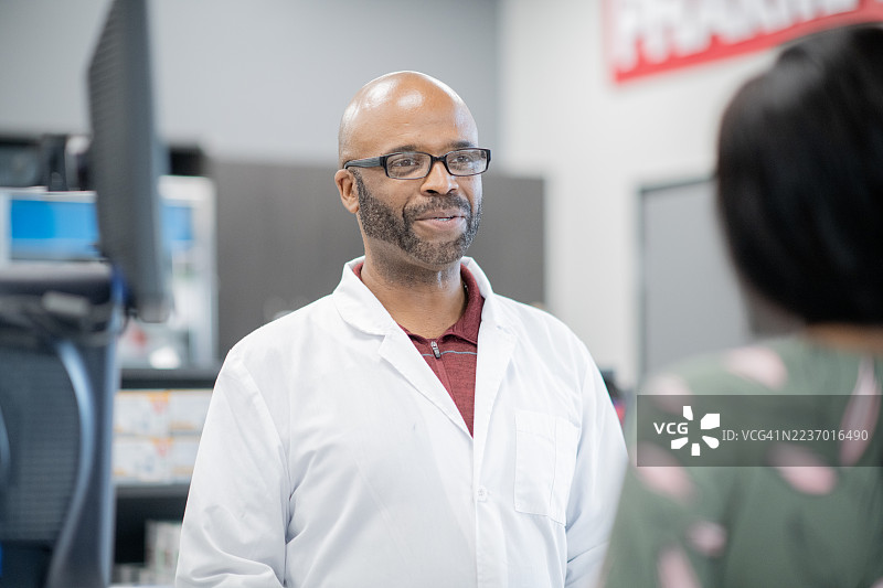 友好的黑人药剂师穿着白色实验室外套，在繁忙的药店环境中与顾客交谈图片素材