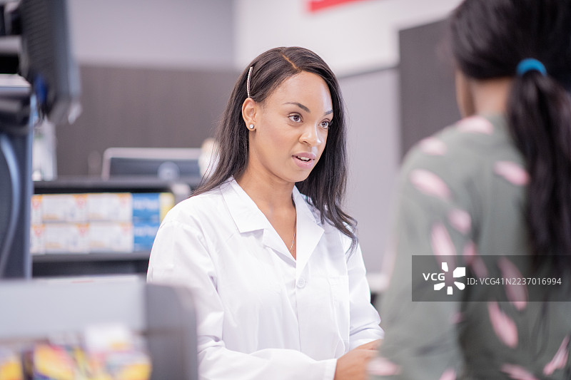 穿着白大褂的药剂师在药房柜台与顾客进行友好的交谈。图片素材