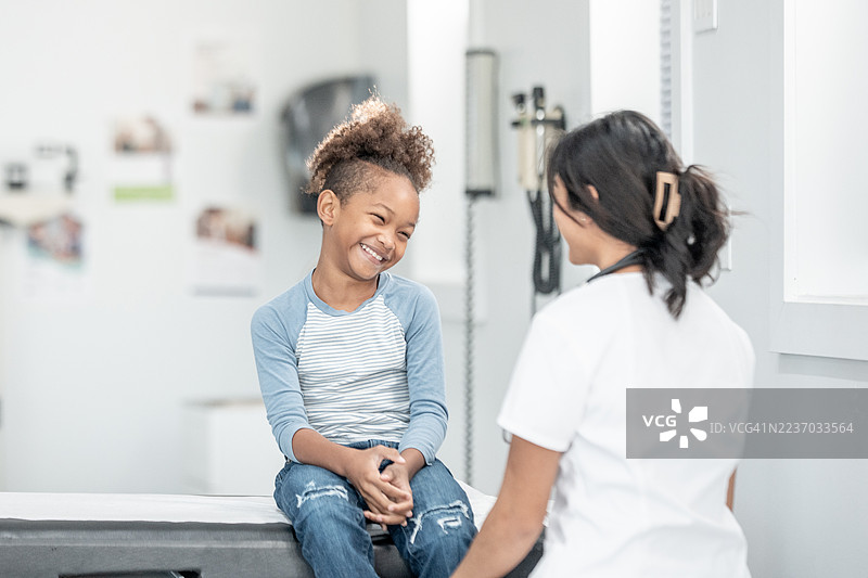 快乐的黑人女孩在明亮的诊所里，在检查台上笑着与护士交谈。图片素材