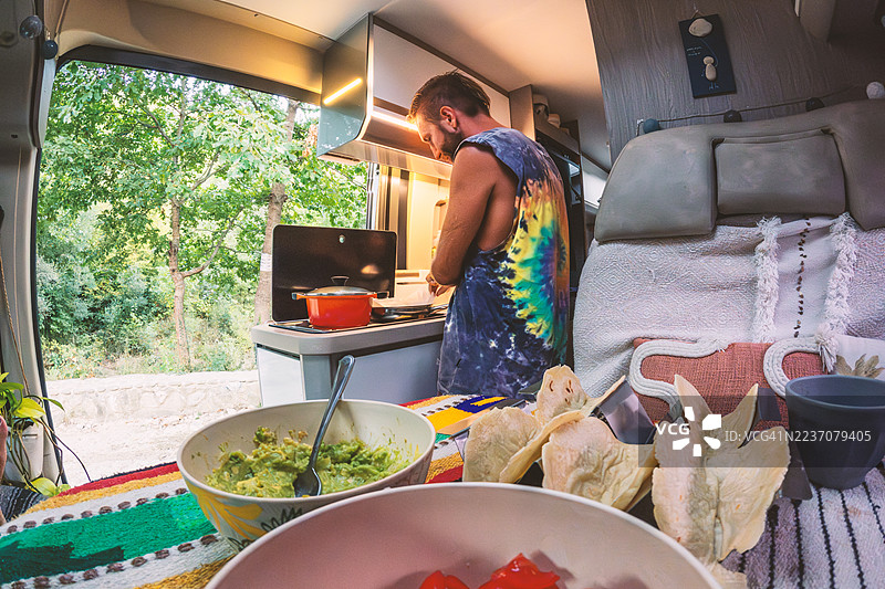 男人在露营车内烹饪墨西哥卷饼，窗外是户外景色图片素材