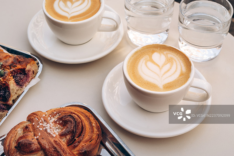 两杯美味的特色卡布奇诺和新鲜出炉的豆蔻面包卷以及巧克力可颂。图片素材