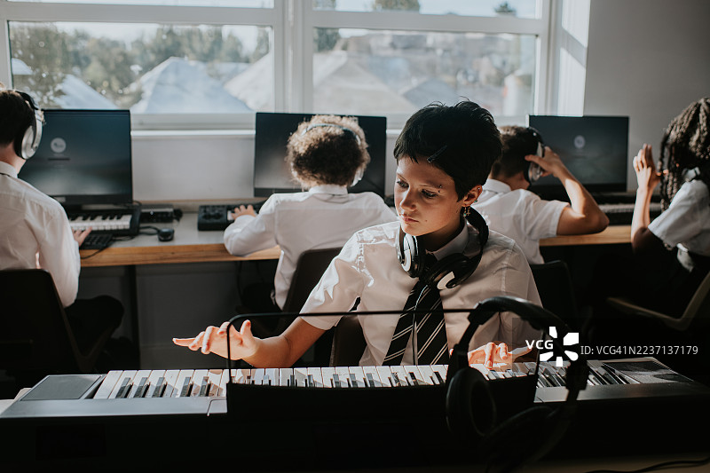 一位另类的年轻音乐学生在教室里弹奏键盘，背景中同学们坐着。图片素材