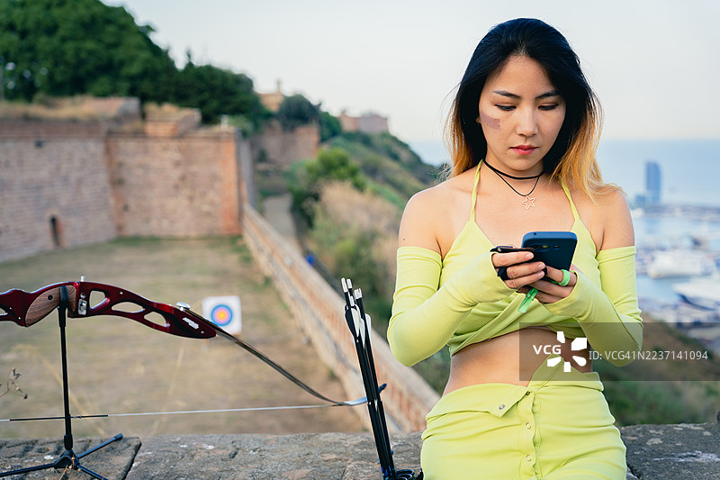 年轻女性在巴塞罗那的蒙特朱伊克城堡练习射箭时查看智能手机，古老与现代休闲活动的结合图片素材