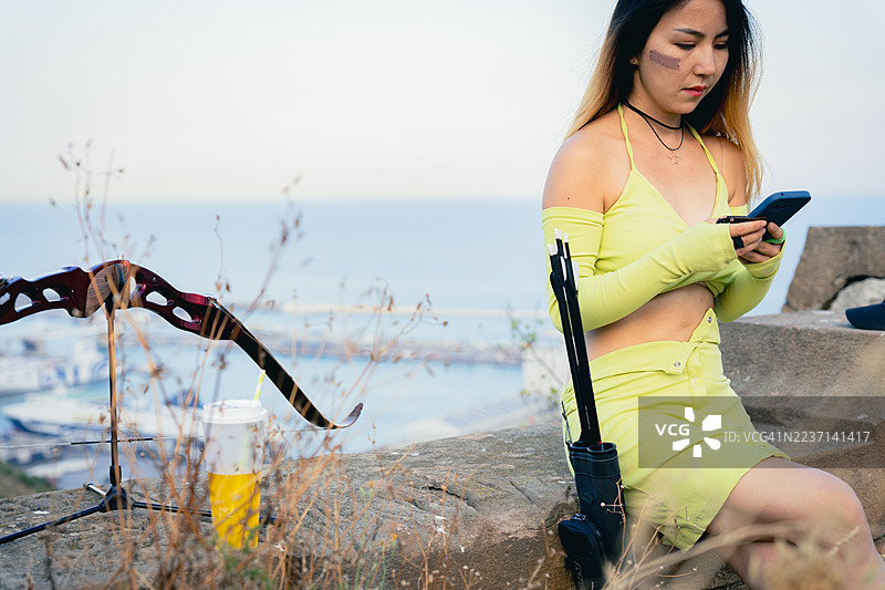 年轻女性弓箭手在海边练习弓箭后查看智能手机图片素材