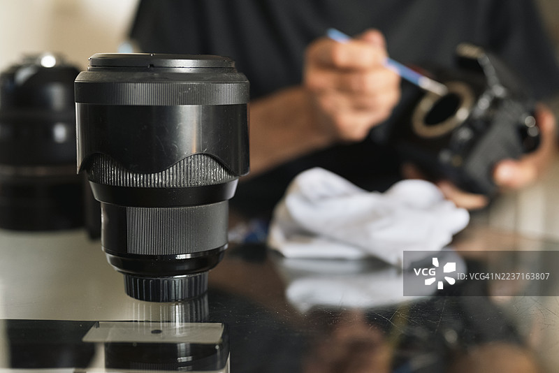 一个男人坐在桌子旁，手中拿着相机，用刷子清洁，前景是相机镜头。图片素材