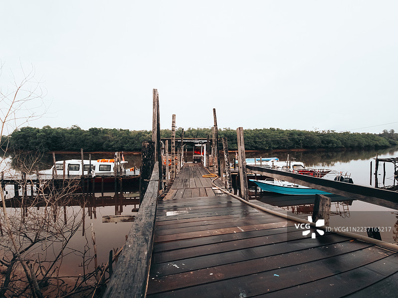 宁静湖泊上的木制码头通往船只，周围环绕着郁郁葱葱的绿树，天空晴朗。马来西亚，美浪（Merang）。图片素材