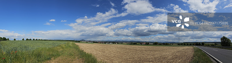 广阔蓝天下，绵绵白云间，一条蜿蜒的道路穿过田野，通向远方的城镇，全景视图。图片素材