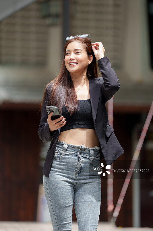 年轻的亚洲女性穿着西装外套和牛仔裤，微笑着拿着手机。图片素材
