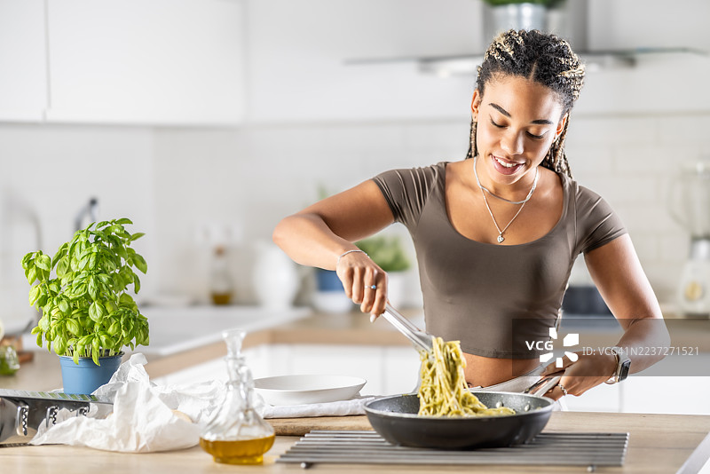 一位聪明的年轻非裔美国女性正在烹饪意大利香草青酱意大利面，她正用勺子搅拌，准备将其盛在盘子里。图片素材