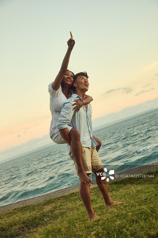 一对情侣在海滩上享受日落，男人扛着女人走，并指向天空。图片素材